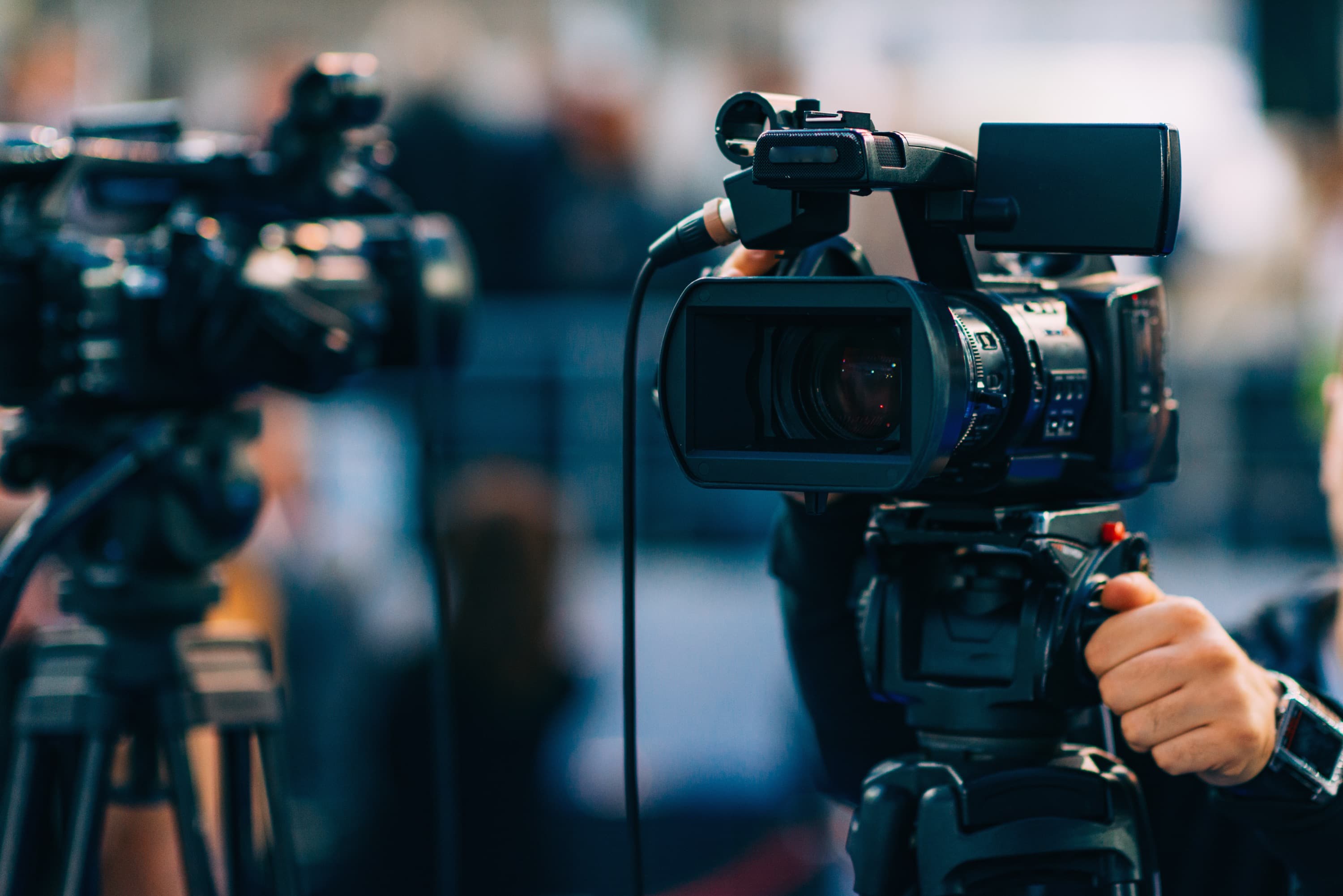 A video camcorder on a tripod, with a camera operator hand on the tripod head and microphone cable extending from the top of the camcorder.