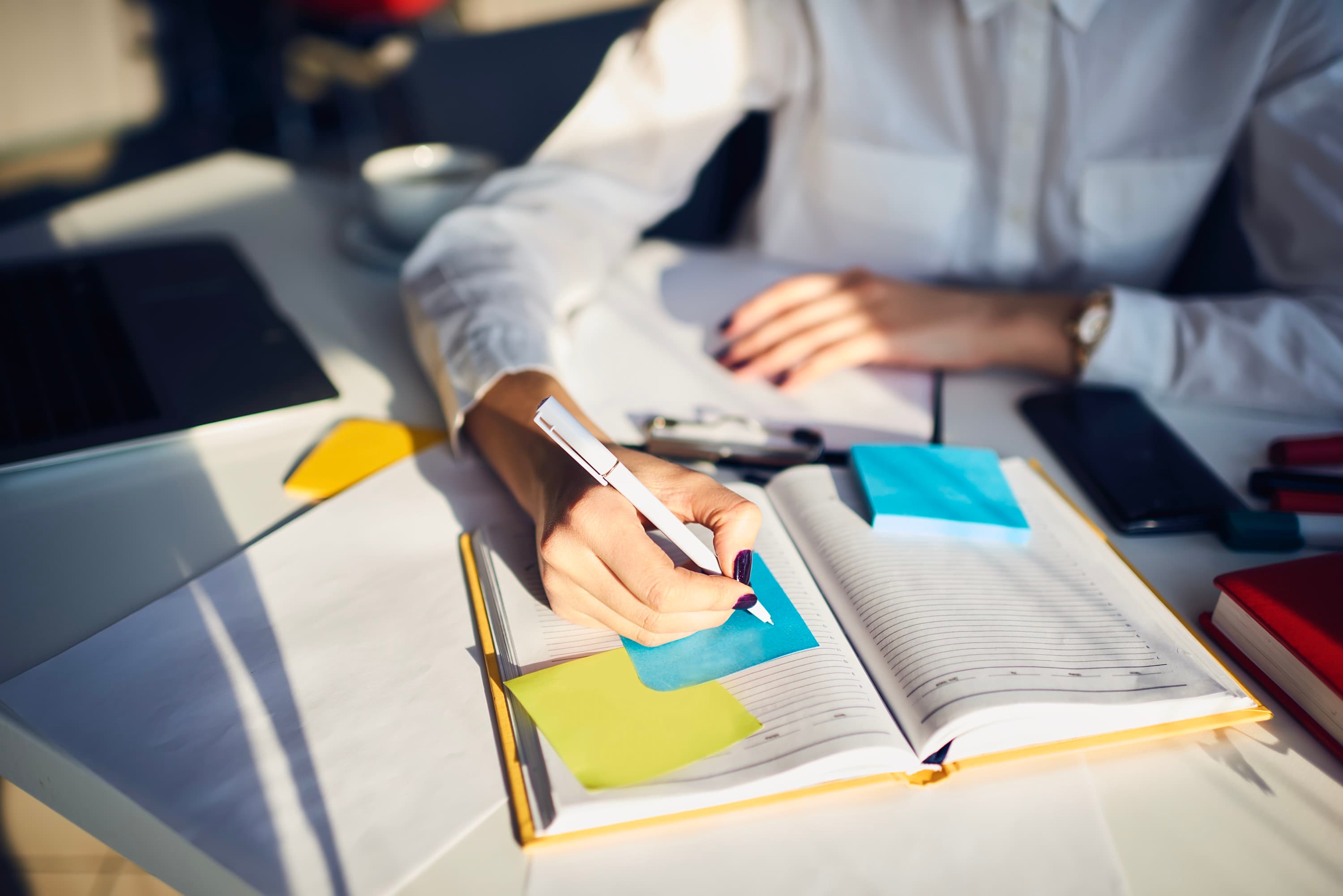 A woman's hand writing on a sticky note in a notepad, with books and other desk items scattered about.